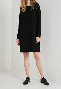 Czarna sukienka z długim rękawem o luźnym kroju, z zamykaną kieszenią po boku. Gładka tekstura i minimalistyczny design.