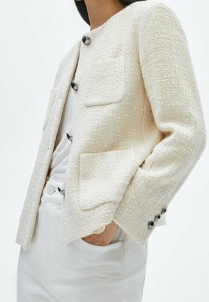 Personne portant une veste crème texturée avec des boutons noirs, des poches plaquées à l'avant et un pantalon blanc, main dans la poche, sur un fond uni.