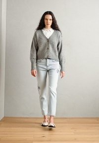 Lys grå strikket cardigan med store knapper, kombinert med lyseblå jeans med blomsterbroderi, og hvite sko med svarte detaljer.