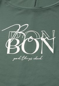 Zelená tkanina s velkým bílým textem, který zní "BON" s stylizovaným „o“ a pod ním menším písmem "dobré věci před námi".