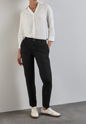 Persona con camisa blanca de manga larga abotonada, pantalones negros y zapatillas blancas, de pie frente a un fondo neutro.