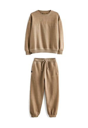 Bež dukserica i hlače od mekog materijala. Dukserica ima urezanu oznaku "SUPERDRY" na prednjoj strani; hlače imaju elastične završetke i remen za stezanje.