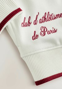 Bílý sportovní top s vroubkovanými manžetami ve tmavě červené barvě a vyšitým textem "club d’athlétisme de Paris" v odpovídající barvě. Hladký povrch.