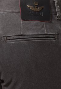 Poche arrière d'un pantalon gris foncé avec une étiquette de marque en cuir noir ornée d'un emblème doré d'aigle et de couronne et des initiales de la marque brodées.