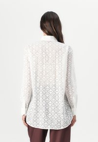 Hvit, gjennomsiktig skjorte med knapper som har et geometrisk mønster i grått. Lange ermer og en buet hemline fremhever den avslappede passformen.