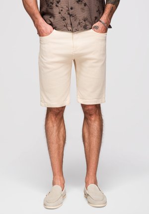 Homme portant un short beige, des chaussures légères beige clair sans lacets, et une chemise marron à manches courtes avec les mains dans les poches, sur un fond uni.