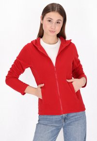 Czerwony polarowy hoodie na zamek błyskawiczny z suwakiem z przodu, ściągaczami przy mankietach oraz dwiema bocznymi kieszeniami. Noszony na białej koszuli i zestawiony z niebieskimi dżinsami.