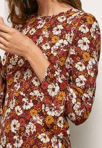 Kvinna som bär en långärmad topp med ett tät blommönster i vitt, gult och rött på en mörkröd bakgrund, hand lyft nära ansiktet.