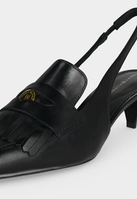Μαύρο δερμάτινο παπούτσι slingback με χαμηλό τακούνι, λεπτομέρεια με φούντες και μικρό χρυσό έμβλημα στο μπροστινό λουράκι.