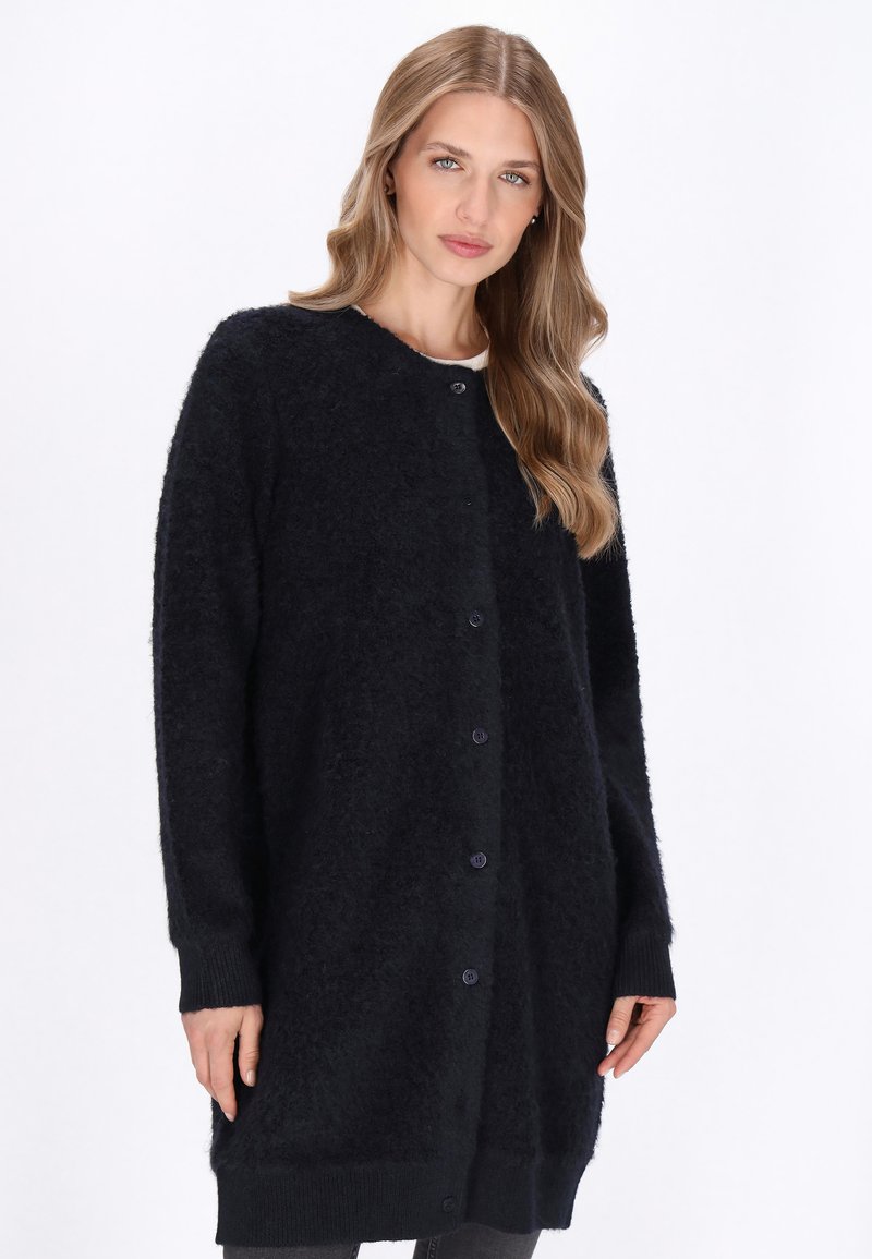 Vrouw met lang blond haar die een lange zwarte fuzzy geknoopte cardigan over een witte top draagt tegen een effen witte achtergrond.