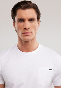 Giovane uomo con capelli castani pettinati, occhi azzurri, espressione neutra, che indossa una maglietta bianca semplice con taschino sul petto e piccola etichetta nera.