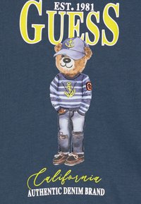 Miś teddy noszący pasek sweter z kotwicą, czapkę, podarte dżinsy i buty żeglarskie pod dużym napisem "GUESS" oraz napisami "California Authentic Denim Brand".