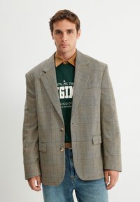 Grau kariertes Blazer mit braunen und weißen Karos, strukturierte Schultern, einzelner Knopfverschluss, über einem grünen Sweatshirt und einem beigen Hemd getragen.