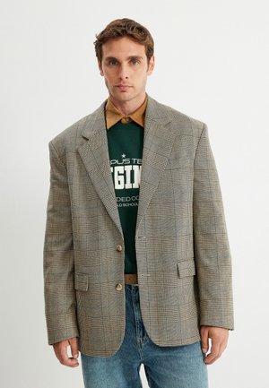 Blazer grigio a quadri con controlli marroni e bianchi, spalle strutturate, chiusura con un solo bottone, indossato sopra una felpa verde e una camicia beige.