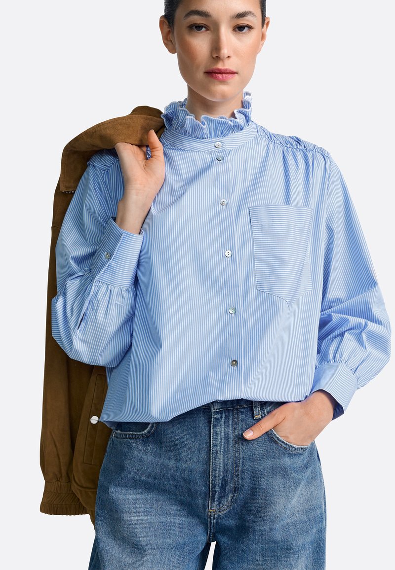 Light blue, striped button-up shirt with a ruffled collar, long puff sleeves, and a front pocket. Paired with high-waisted denim jeans.