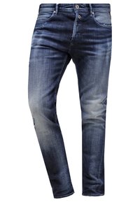 Jean slim bleu foncé délavé avec des effets de blanchiment et une légère usure, avec poches avant, passants pour ceinture et fermeture par bouton.