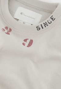 Felpa beige chiaro in tessuto strutturato. Presenta accenti stampati in scuro e rosso sul colletto, inclusa la parola "SINCE". Etichetta visibile.