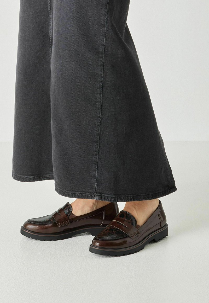 Mocassins en cuir marron avec un détail noir texturé portés avec un pantalon noir à jambes larges sur un sol blanc.