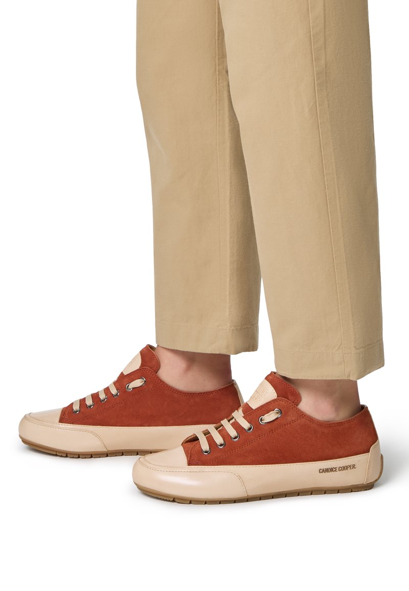 Des baskets en daim rouge présentent des accents et des lacets en cuir beige, avec une semelle en caoutchouc et un logo embossé. Portées avec un pantalon court de couleur claire.