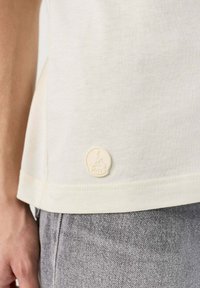 Camiseta de color crema claro hecha de una tela suave, con un parche redondo del logo de JOTT en el dobladillo. Jeans de denim gris con una textura sutil visible.