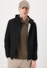 Sort jakke med lynlåslukning, hætte og elastiske ærmer over en brun struktureret sweater, båret med en beige baseballkasket.