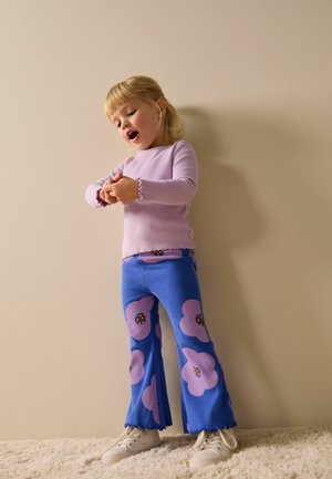 Lyse lilla langærmet top med bølget kant, kombineret med blå vidde bukser med store pink blomster og mørkebrune midter. Hvide sneakers.