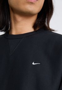 Pogled od blizu na vrat in brado osebe, ki nosi črno sudbrr sweatshirt s belim logotipom Nike na prsih, dolgi temni lasje.