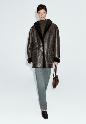 Donna che indossa un cappotto in pelle marrone scuro oversize con rifiniture in pelliccia, jeans grigi, scarpe nere, mentre tiene una borsa marrone su uno sfondo bianco semplice.