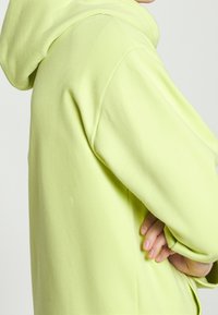 Sudadera verde claro de tela suave, con una gran capucha y un corte holgado. La manga presenta una textura suave con costuras mínimas.