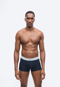 Αθλητικός άντρας που στέκεται με το πρόσωπο προς τα εμπρός, φορώντας μαύρα κοντά εσώρουχα Tommy Hilfiger με λευκή μέση, μπροστά σε απλό ανοιχτό φόντο.