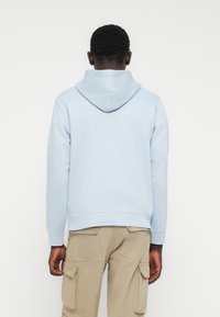 Mężczyzna stojący tyłem, ubrany w jasnoniebieską bluzę z kapturem oraz beżowe spodnie typu cargo z tylnymi i bocznymi kieszeniami.