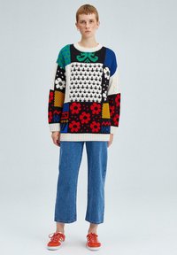 Pull tricoté coloré avec divers motifs, incluant des fleurs et des dessins géométriques. Associé à un pantalon court en denim bleu et des baskets rouges.