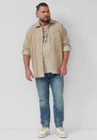 Beige corduroy shirt over een lichtgrijs T-shirt, gecombineerd met lichtblauwe jeans en witte sneakers. Casual fit met opgerolde mouwen.
