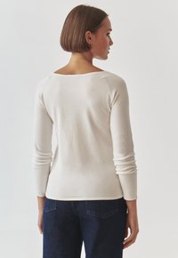 Weißes Langarmoberteil mit Rundhalsausschnitt, taillierte Silhouette und gerippten Bündchen, kombiniert mit dunkelblauen Denim-Jeans. Glatte Stofftextur.