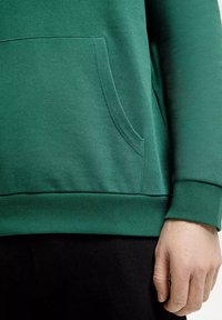 Zielona bluza z kangurzą kieszenią, ściągaczami przy mankietach i u dołu, wykonana z miękkiego materiału o gładkiej fakturze.