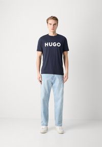T-shirt din bumbac navy cu inscripția "HUGO" în litere albe, asortat cu blugi ușori albaștri și adidași albi. Modelul stă pe un fundal simplu.