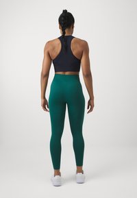Black sports bra with racerback design, paired with high-waisted green leggings. The model wears white athletic shoes.