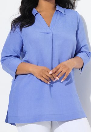 Femme portant un chemisier tunique bleu clair à manches longues avec un col en V, les mains doucement jointes devant, associée à un pantalon blanc.