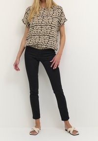 Top beige con patrón abstracto en negro, de mangas cortas, combinado con jeans ajustados negros y sandalias de tira crema con detalle de nudo.