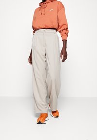 Orange hoodie med framficka och vitt logotyp, parat med beige veckade vida byxor och orangea och vita sneakers.