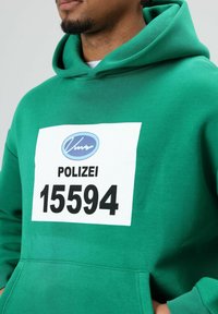 Groene hoodie met een grote witte patch waarop in het zwart de tekst "POLIZEI 15594" staat, met een gladde textuur en een klassieke pasvorm.