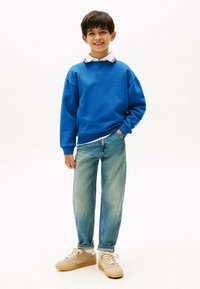 Sudadera de algod�ón azul con puños acanalados, combinada con jeans de mezclilla de lavado claro. Zapatillas beige con cordones blancos. Camisa con cuello blanca y roja debajo.