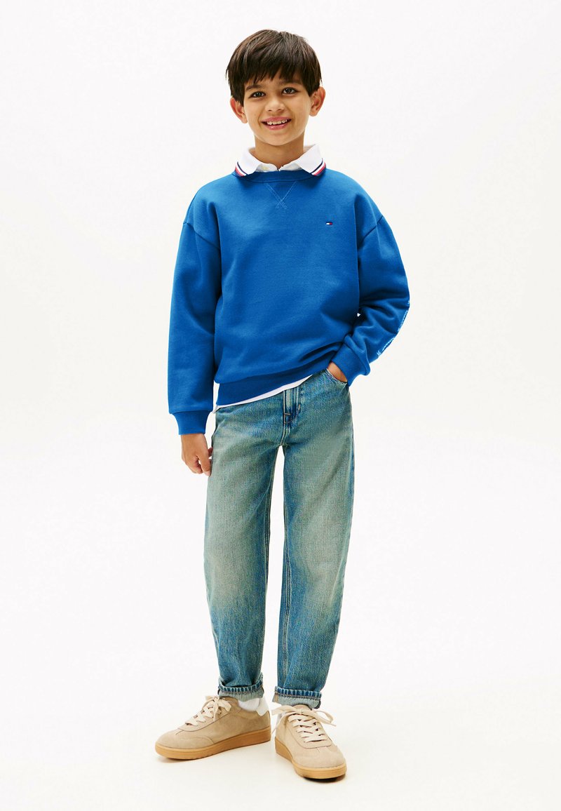 Sudadera de algodón azul con puños acanalados, combinada con jeans de mezclilla de lavado claro. Zapatillas beige con cordones blancos. Camisa con cuello blanca y roja debajo.