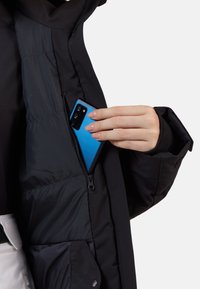 Czarna kurtka puchowa z zamykaną kieszenią wewnętrzną. Niebieski smartfon jest wkładany do środka. Materiał wygląda na gładki i ocieplany.