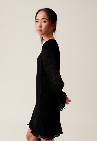 Robe noire plissée avec de longues manches fluides et un ourlet volanté, mettant en valeur une texture lisse et une silhouette élégante sur un fond neutre.