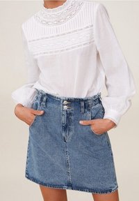 Blouse blanche avec des détails en dentelle complexe et des plis, associée à une jupe en denim bleu clair à coupe décontractée et deux poches avant.