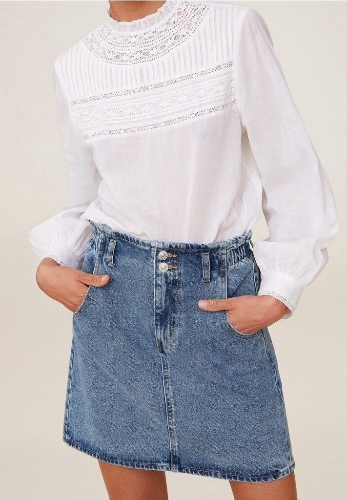 Blouse blanche avec des détails en dentelle complexe et des plis, associée à une jupe en denim bleu clair à coupe décontractée et deux poches avant.