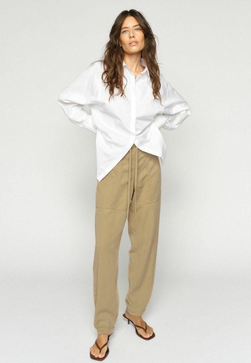 Mujer con cabello largo y ondulado, vestida con una camisa blanca de gran tamaño, pantalones beige con cordón y sandalias marrones de tacón, de pie con las manos en las caderas.