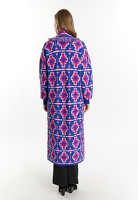 Palton lung, tricotat, cu un model geometric îndrăzneț în nuanțe de albastru, roz și alb. Designul include un guler ciufulit și mâneci lungi.