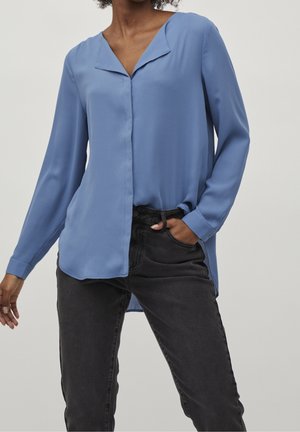 Vrouw draagt een lichtblauwe blouse met lange mouwen, lichtjes ingestopt in een hooggetailleerde zwarte spijkerbroek, met één hand in de zak.